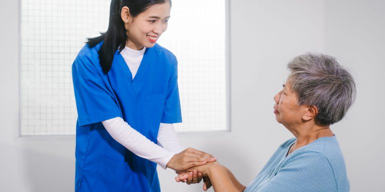An Asian female nurse provides caring home care to an elderly woman, assisting with arm movement